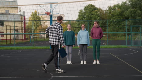 jugadores de voleibol principiantes de pie en la cancha al aire libre, sonriendo mientras se preparan para la sesión de entrenamiento con el entrenador, la cancha deportiva está rodeada de árboles, una valla metálica y edificios residenciales