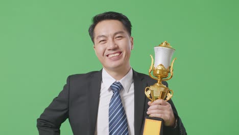 close up of asian business man in a suit and tie with trophy smiling to camera with one arm akimbo posing as the first winner on green screen background in the studio