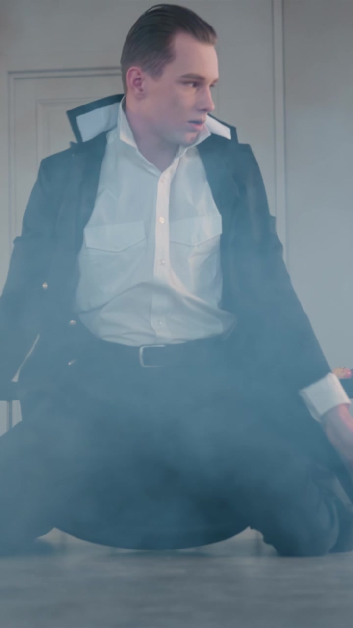 Giovane uomo in abito formale che assume varie posture. L'uomo interagisce con i suoi vestiti, mentre sono presenti piccoli tavoli. L'ambientazione presenta una semplice porta bianca, con il giovane uomo in evidenza.