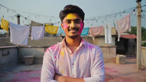happy young man celebrating holi festival on a rooftop with arms crossed, smiling with colored powder on his face and clothes, and colorful fabrics hanging in the background