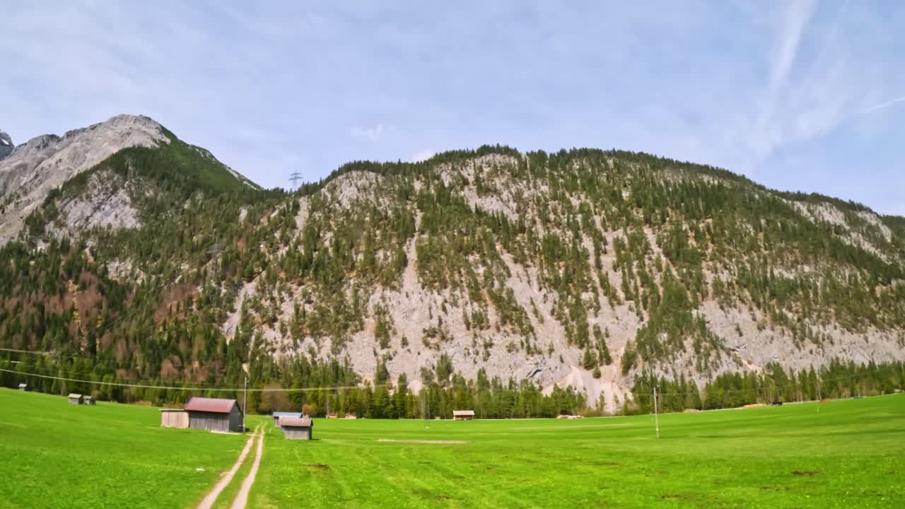 Green meadow punctuated by clustered farm sheds spreads below a steep forested limestone slope in the Loisachtal near Garmisch-Partenkirchen, along the Innsbruck–Munich train in the Bavarian Alps