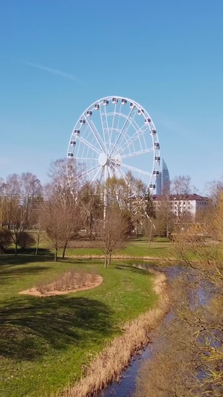Ferris wheel near river in Riga with blue sky and distant business buildings