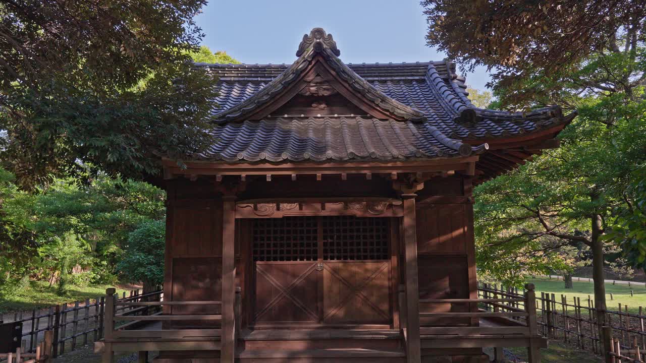 A traditional Japanese wooden shrine with an ornate roof is nestled among green trees in a park setting, under a clear sky.