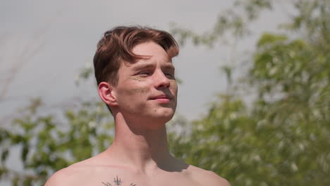 shirtless young man loosening headband pushing head back eyes closed inhaling fresh breeze by riverside surrounded by green grasses trees tattooed chest under bright daylight outdoors adventure