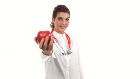 young friendly female nutritionist showing a red apple, advising healthy diet. portrait of young professional with stethoscope and lab coat isolated on white background