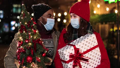vista de cerca de una feliz pareja multiétnica con máscaras faciales hablando y sosteniendo regalos de navidad en la calle mientras nieva en navidad