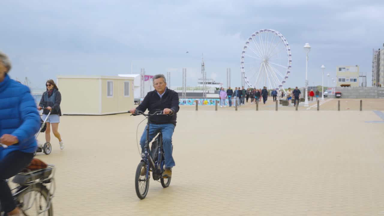 People walking on sea promenade near the beach of Nieuwpoort with ferris wheel in background. Holiday town by the Belgian sea - Slow motion