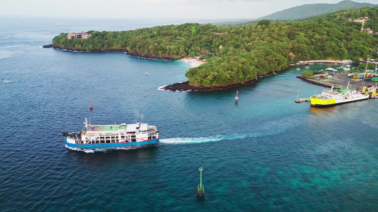 Porto di un'isola tropicale con un traghetto in partenza attraverso acque calme e blu, circondato dalla costa e dal terminal dei traghetti a Padang Bai, Bali, Indonesia, filmato drone con ripresa in allontanamento.
