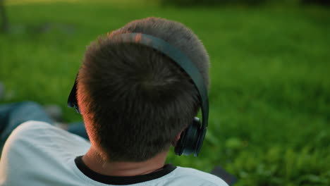 back view of music producer wearing headphones, nodding to music while operating phone, seated on grassy field with sunlight reflecting off his head