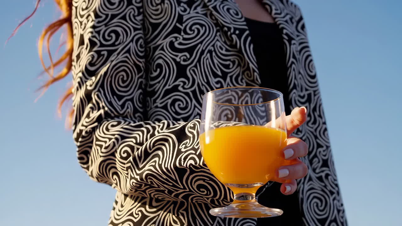 Person in a patterned jacket holding a glass of orange juice outdoors