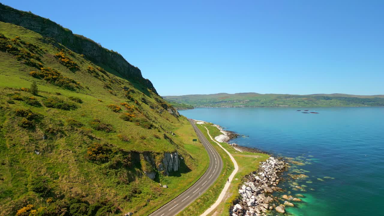Low, wide aerial video of a snaking road on the Causeway Coastal Route in County Antrim, Northern Ireland, UK, on a bright and sunny day. Filmed in 4K, 60FPS and with Rec709 color.