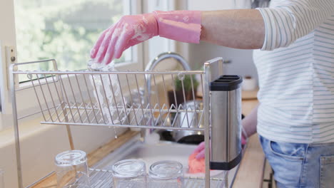 Middle-aged-hands-of-caucasian-woman-washing-up-in-gloves-in-kitchen-with-copy-space