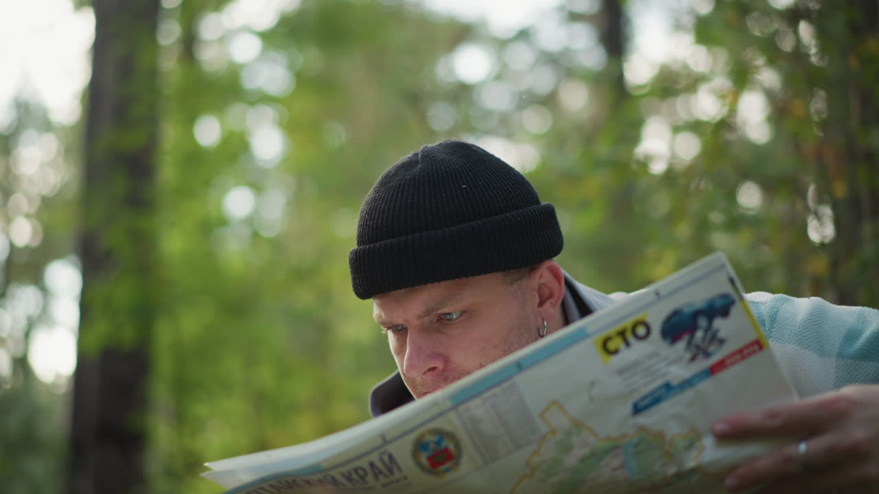 white man closeup reading map, narrows eyes in forest, studies route details, squints at markings, tilts head to trace trail, adjusts beanie, thoughtful expression, dappled sunlight, intimate portrait