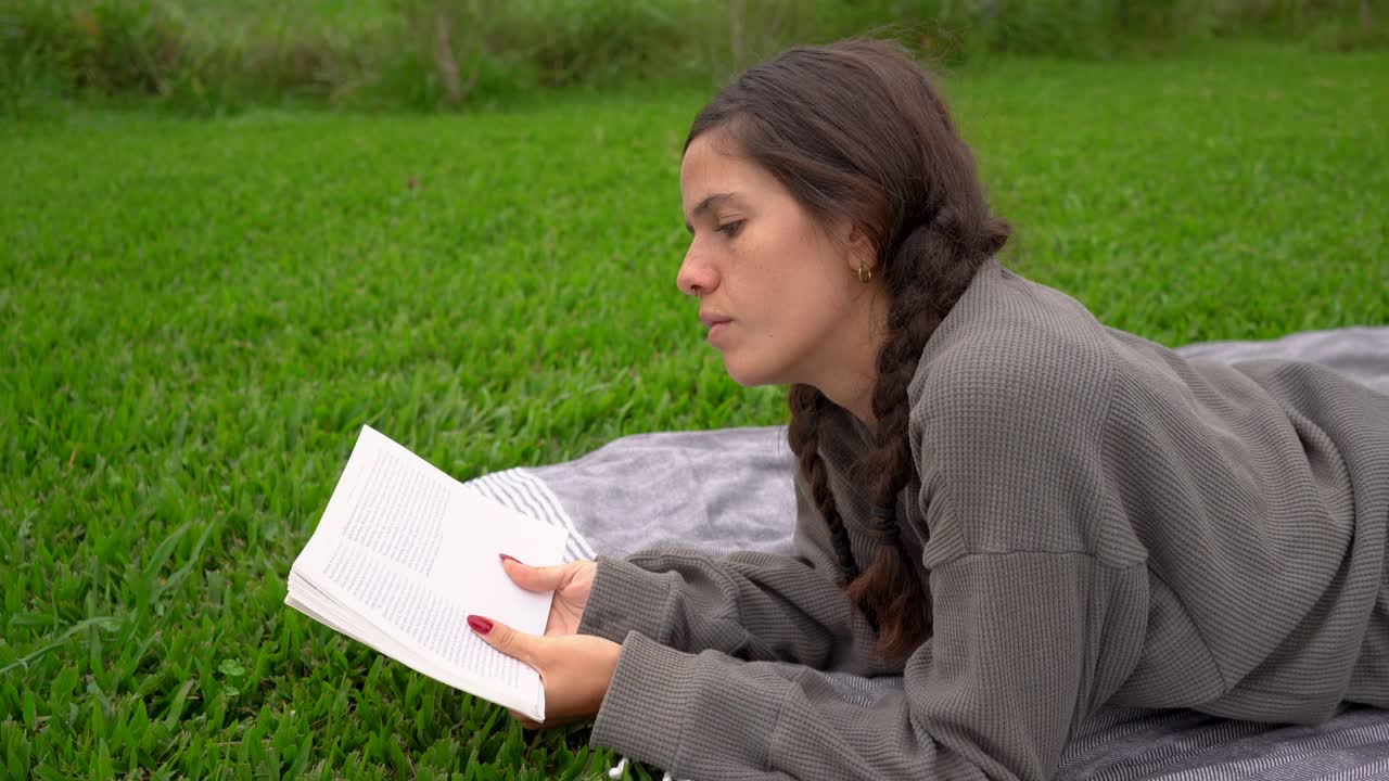 Giovane donna bruna sdraiata su una coperta sul prato che legge un libro, vista laterale a mezzo busto