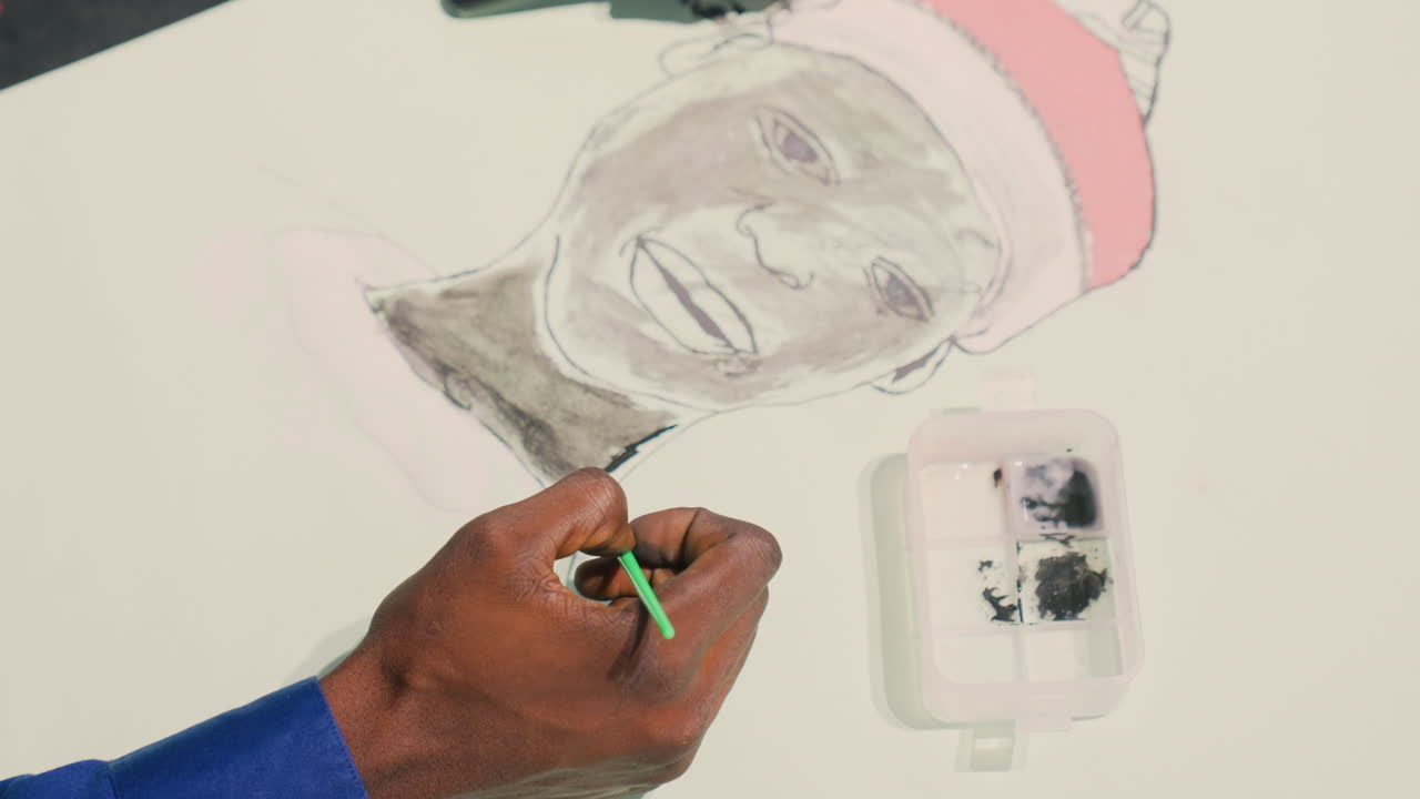 Black painting portrait with green brush, Black illustrator adds fine watercolor highlights to cheek and beanie, closeup of darkskinned hands holding small brush over palette tray, gentle strokes