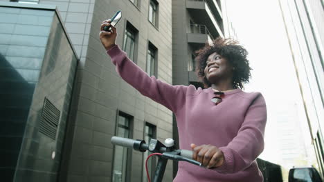 cheerful african american woman standing in city with e-scooter and taking selfie