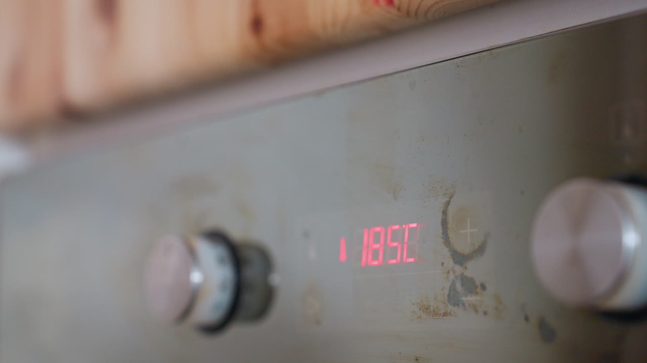 Close up of kitchen digital oven control panel displaying 185C with glowing red LED temperature setting, stainless steel surface, and visible knobs and smudges showing real-life cooking usage