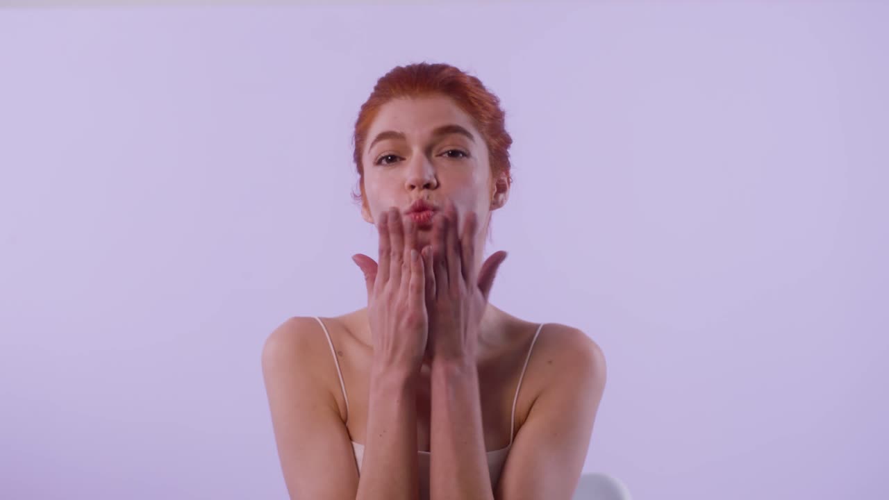 A red-haired woman moves her head side to side while touching her cheeks, then blows a kiss and exits the scene with a smile.