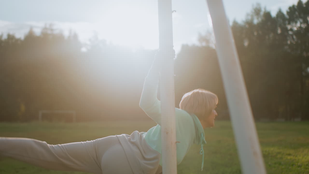 Graceful woman performing waist stretch exercise outdoors holding metal pole under bright sunlight on grassy field in peaceful park surrounded by trees demonstrating strength, and healthy lifestyle