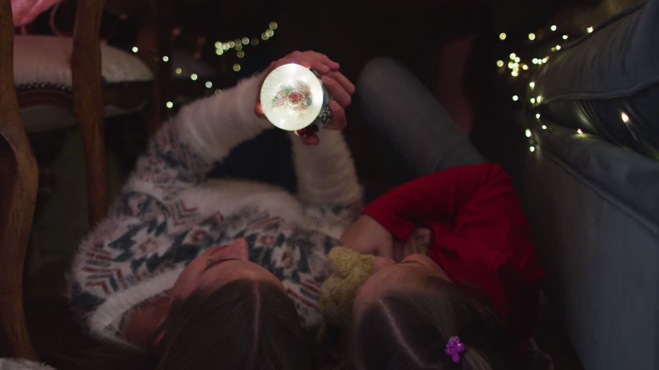 백인 어머니와 딸은 크리스마스 동안 담요 요새 에 누워있는 동안 눈 구를 들고 있습니다.