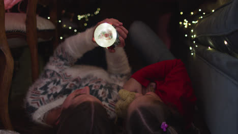 madre y hija caucásicas sosteniendo un globo de nieve mientras yacían bajo una manta fuerte durante la navidad en