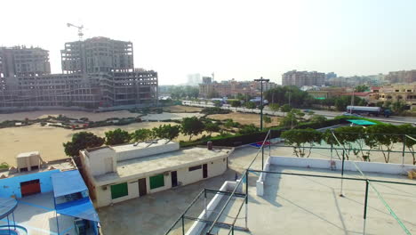 Aerial-tilting-down-view-of-a-highway-of-the-city-with-traffic-going-on,-under-constructed-building-with-crane-of-its-top,-a-sports-area-and-empty-plot-with-its-roads