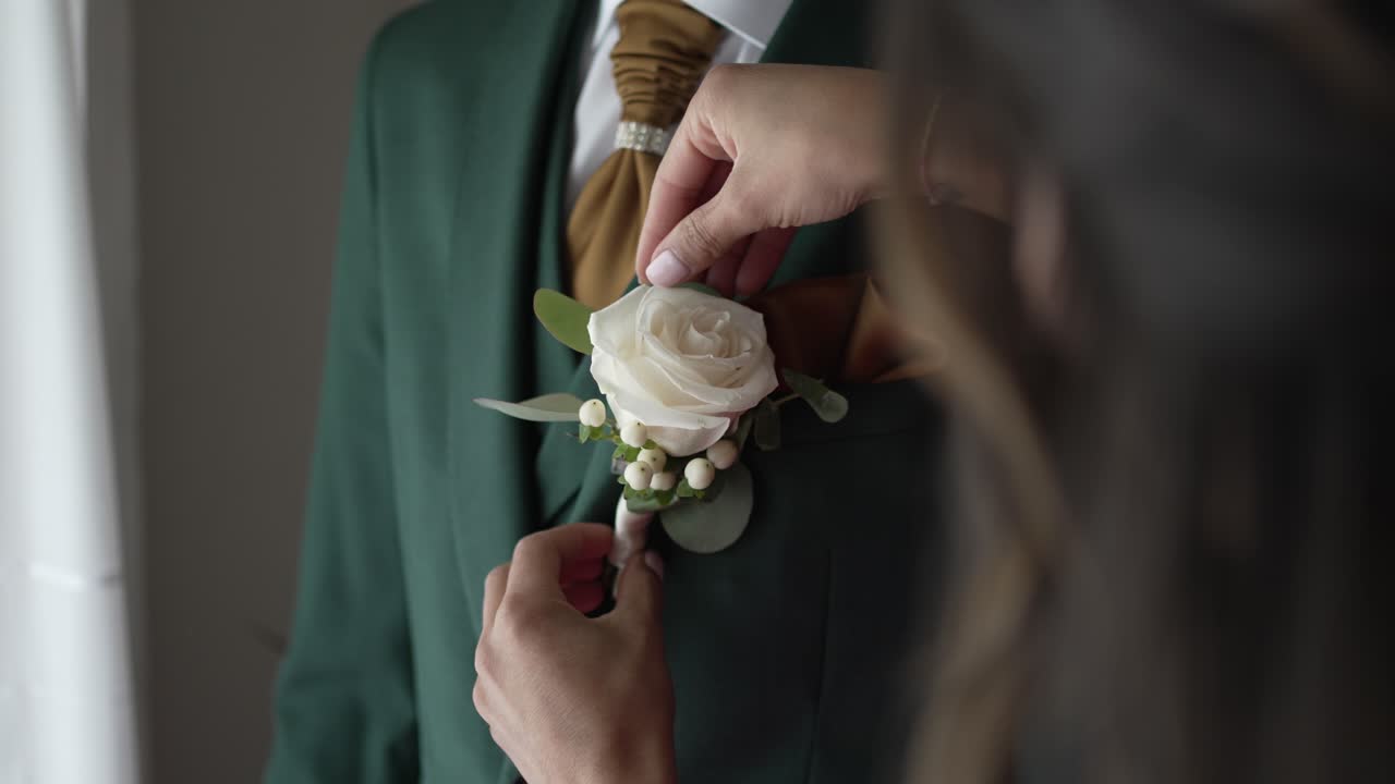 Close up of bride pinning white rose boutonniere on groom’s green suit before wedding