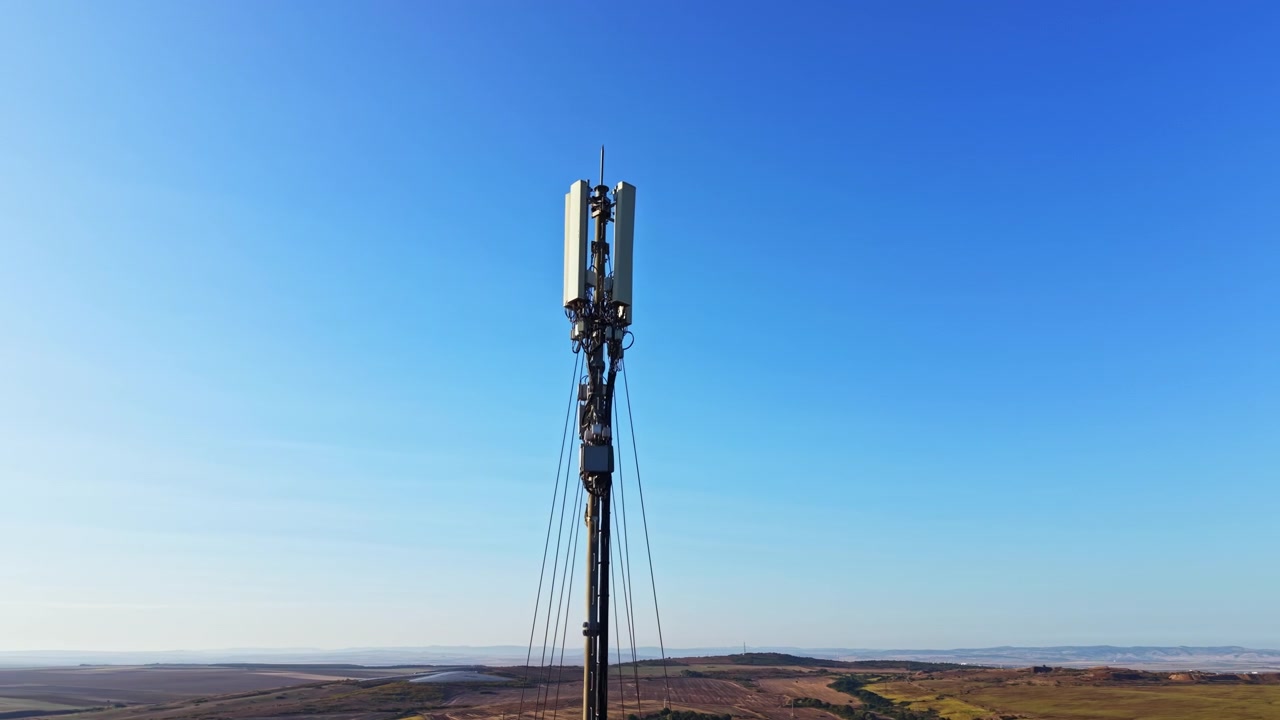 Una torre cellulare si erge alta contro un cielo azzurro e limpido in un vasto paesaggio aperto con dolci colline e campi sotto la luce del sole.