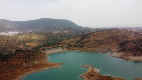 Acercamiento-Aéreo-Al-Lago-Azul-De-La-Presa-Hidráulica-Rodeado-De-Hierba-Seca-Y-Montañas