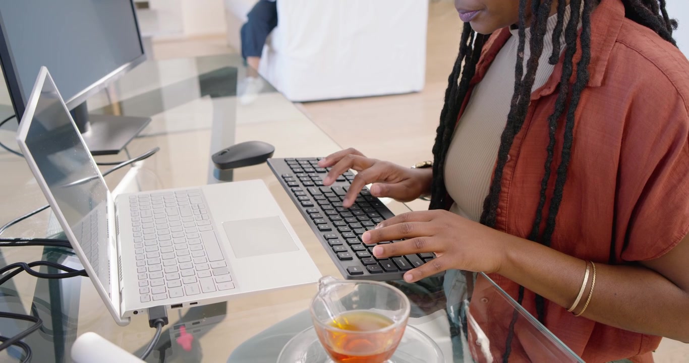 Adult African woman typing on black keyboard, using mouse, working on white laptop with teacup