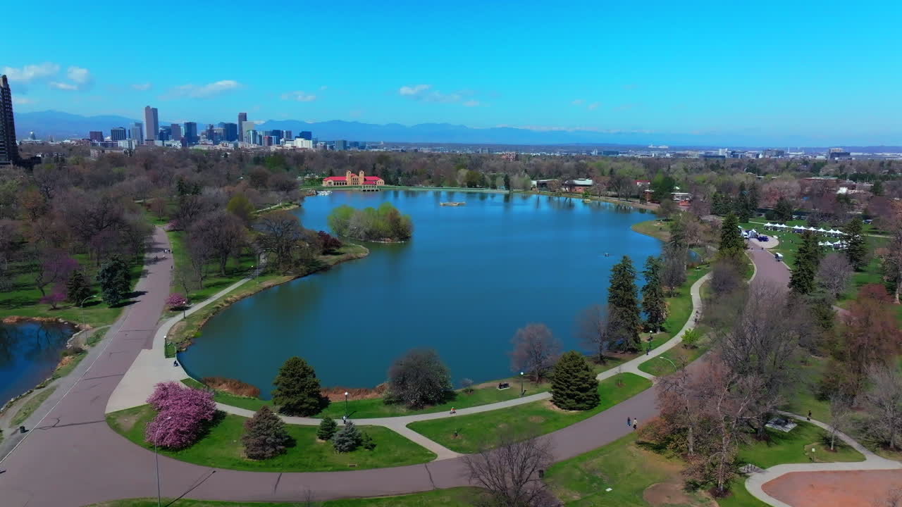 Springtime City Park Denver Colorado Lake Pavilion vibrant spring summer aerial drone sunny blue sky snow capped Rocky Mountains front range cityscape green lush grass trees circle right motion