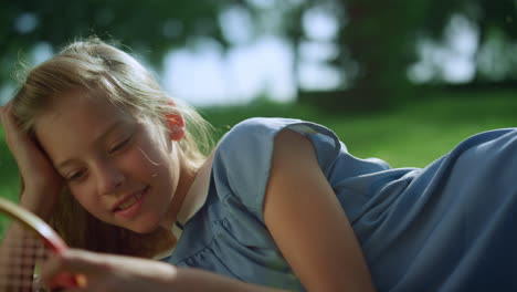 Smiling-girl-lying-on-green-lawn-alone.-Blond-kid-touch-racket-net-closeup