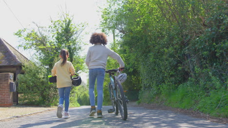 Rückansicht-Eines-Jungen-Mit-Fahrrad-Und-Eines-Mädchens-Mit-Skateboard,-Die-Gemeinsam-Die-Landstraße-Entlang-Gehen