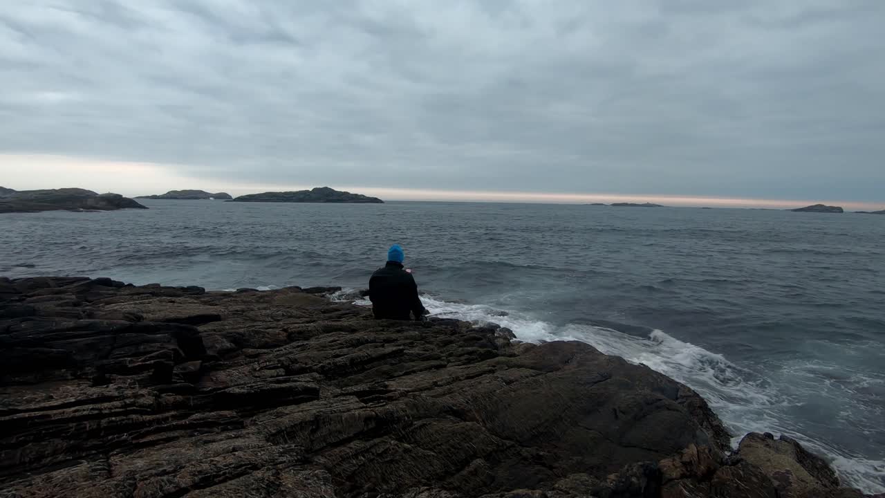 Sad male person sitting alone and using his phone close to sea at coastal rock while waves are hitting - Static evening clip with dramatic clouds after sunset