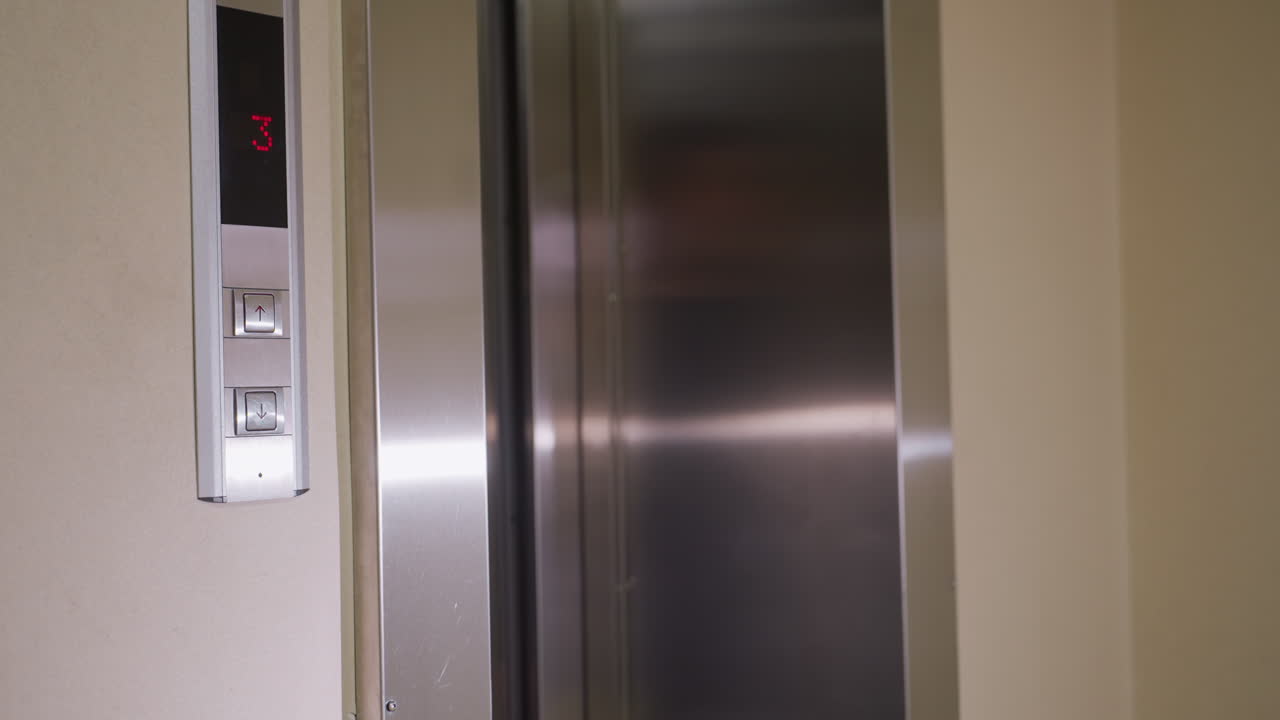 Elevator control panel with up and down arrows, digital display showing floor 3 in red, modern metallic finish with sleek design. Display indicates elevator movement in office building or high-rise