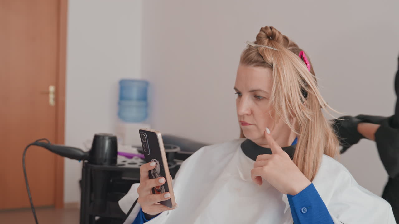 blonde client scrolling phone during salon treatment, stylist prepping foils and dryer while woman sits in white cape, focused expression, modern interior with water cooler, closeup on hands