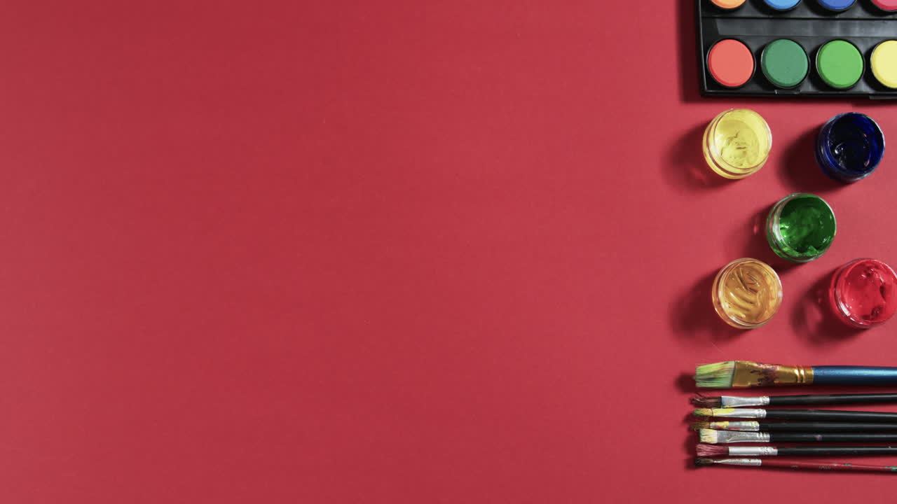 vista aérea de pinturas y pinceles dispuestos sobre un fondo rojo con espacio de copia, en cámara lenta