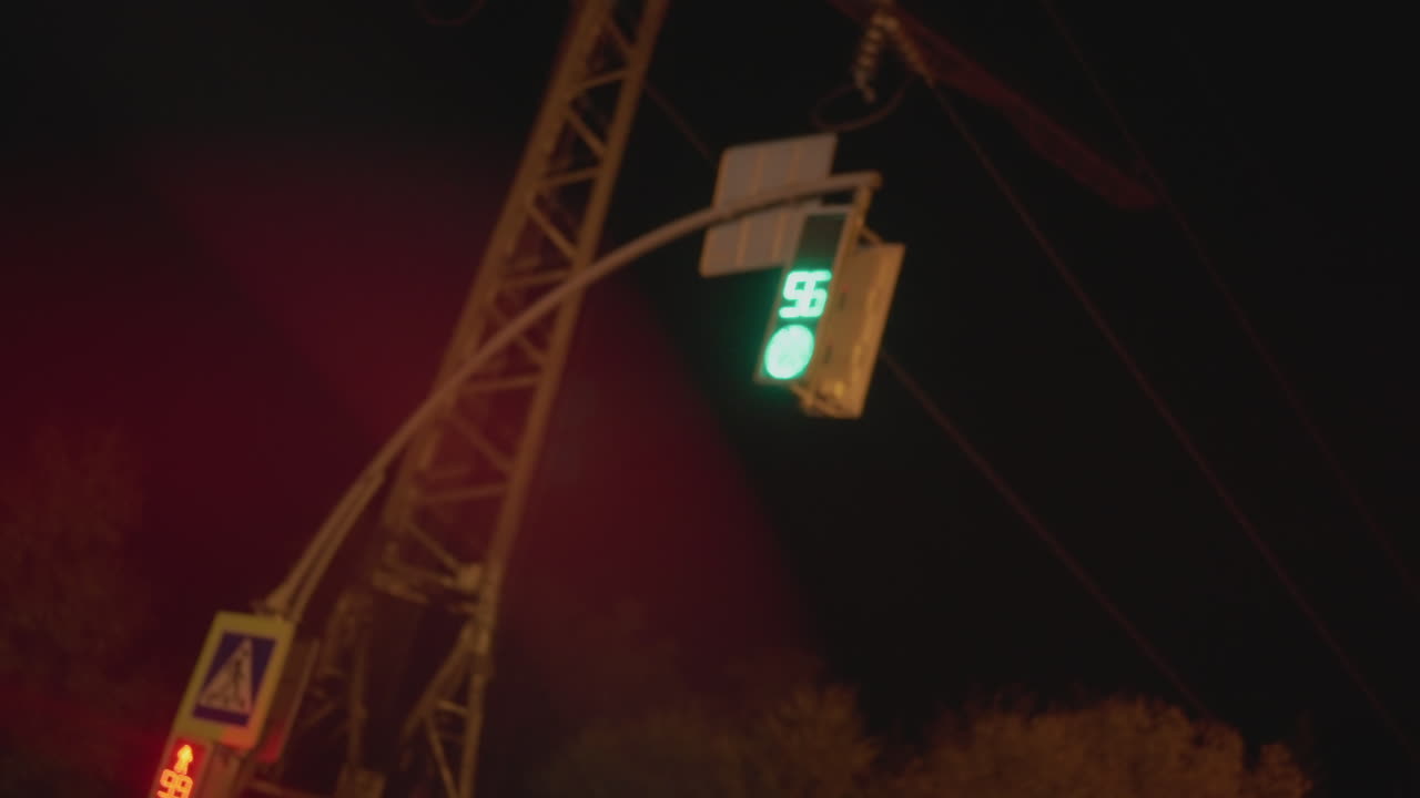 Semáforo verde nocturno colgando sobre una intersección, señal de peatones iluminada y mástil de soporte inclinado, líneas eléctricas cortando el cielo oscuro con destellos rojos en la lente, copas de árboles distantes y sutiles