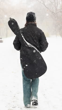 vista trasera de una mujer llevando una guitarra en el caso, caminando en un parque nevado, con copos de nieve aferrados a su chaqueta y cabello, árboles borrosos en el fondo