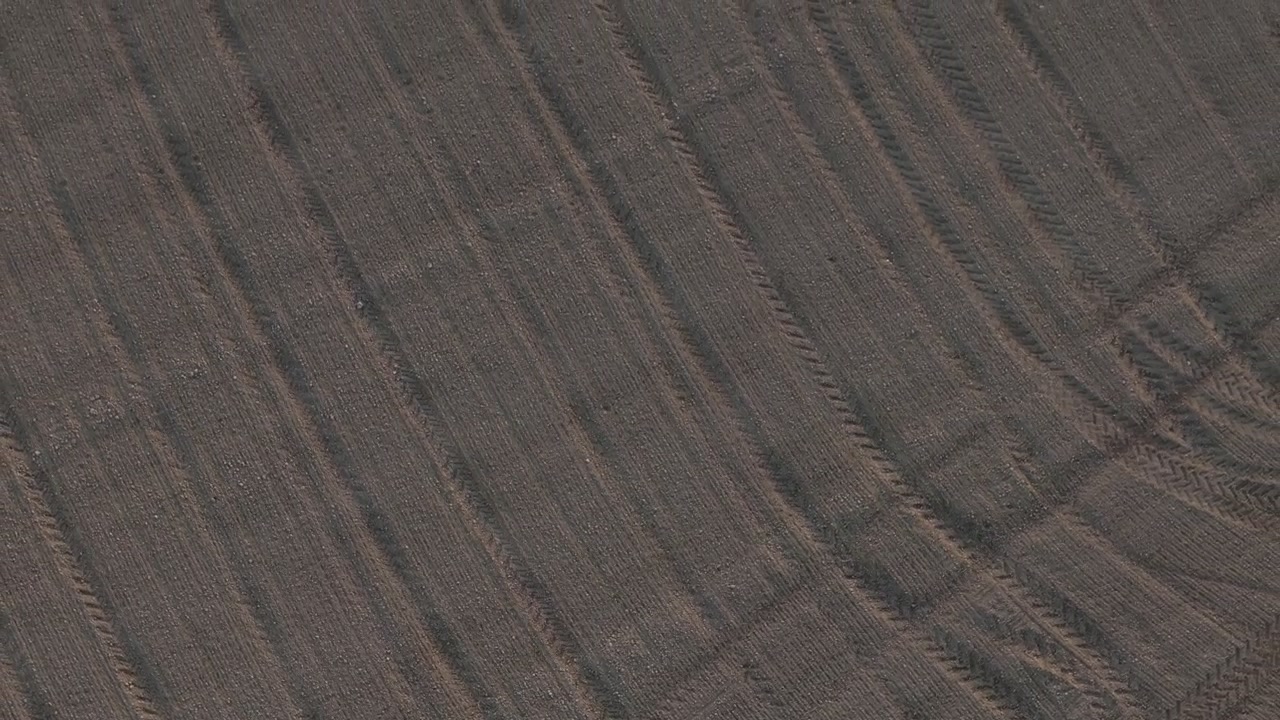 Cultivated farmland in the Po Valley, Italy, revealing mechanized tillage textures, tractor cultivation marks, and dry alluvial soil patterns, ascending drone shot.