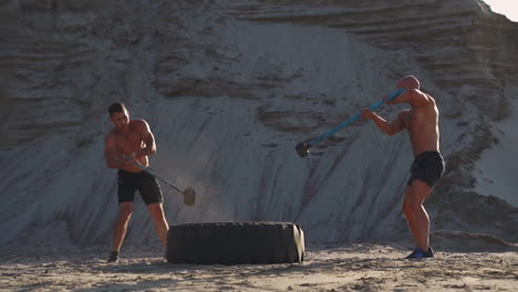 two powerful male athletes training together hit the wheel with a hammer at sunset in the desert. endurance training