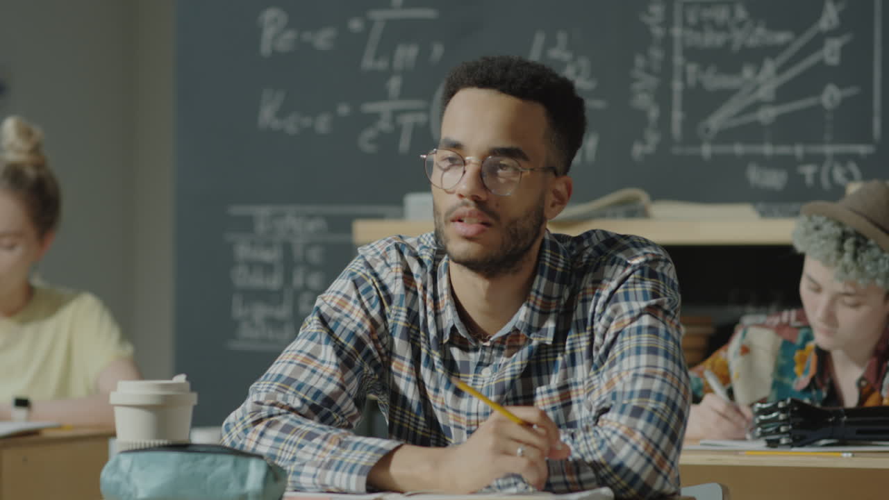 A male student with glasses in a plaid shirt sits at a desk in a classroom, looking thoughtful or engaged, with a blackboard in the background