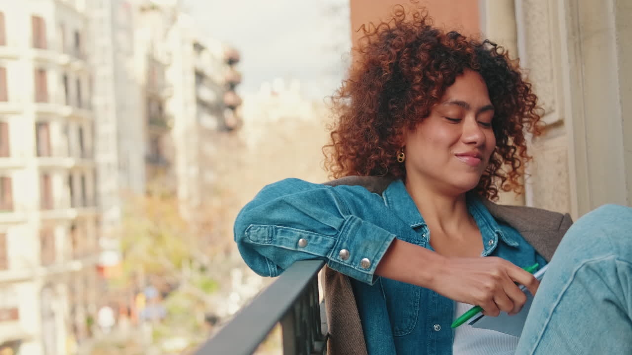 Smiling Young Woman with Curly Hair Writing in Notebook on Balcony
