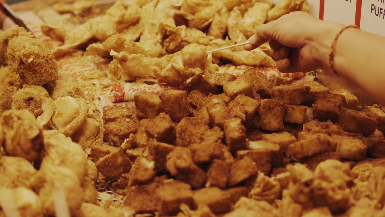 Una vista ravvicinata e dall'alto di una varietà di snack fritti, inclusi tofu fritto dorato e involtini primavera, esposti presso una vivace bancarella di street food in un mercato notturno
