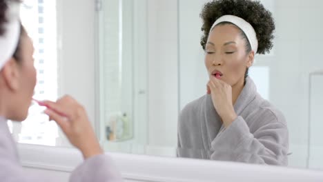 Biracial-woman-in-bathrobe-brushing-teeth-in-bathroom,-slow-motion