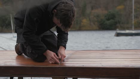 young carpenter uses knife to cut old sealant from in between boat cabin roof deck planks