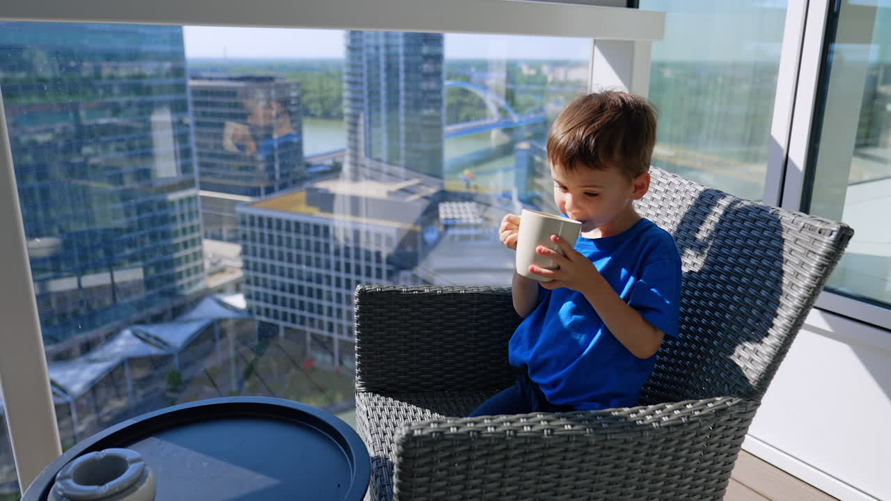 Little kid sitting at the balcony takes a cup from the table. Baby boy drinks from a cup squinting from the bright sun