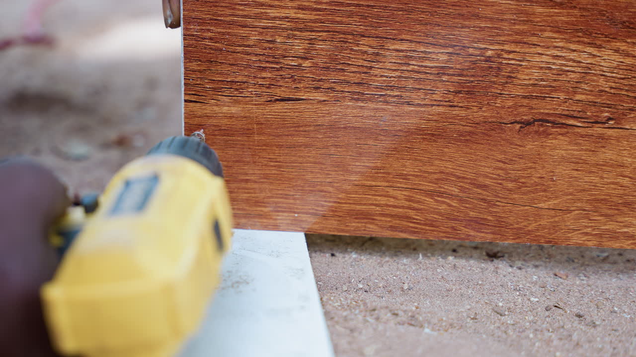 Falegname di colore che fora il bordo di una tavola all'aperto, primo piano di un trapano a batteria che avvita una vite in una tavola stagionata, foro pilota preciso, terreno sabbioso sotto i piedi, scena di installazione di un decking, mani ferme, pratico