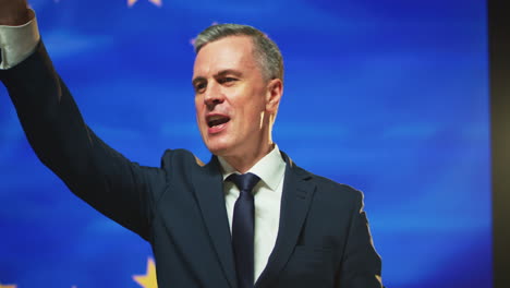 successful politician giving promises against european flag pan around view of man in suit gesticulating and persuading audience during speech against flag of europe on stage
