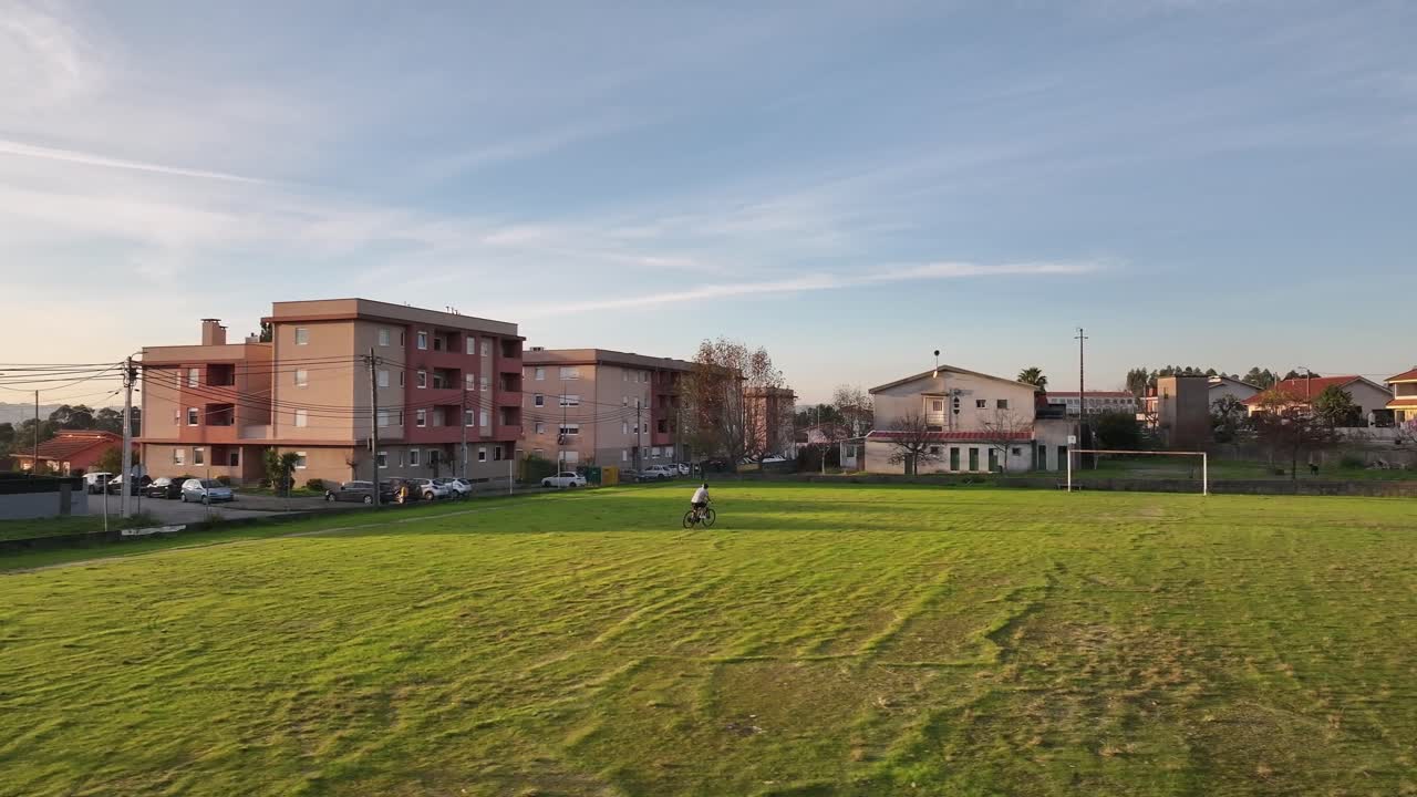 Aerial drone shot of a cyclist riding around a goalpost in a grassy field, revealing a wide open football field football at sunset.